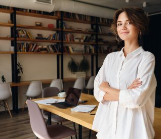 Young attractive woman in white shirt dreamily looking in camera with desk on background in modern office